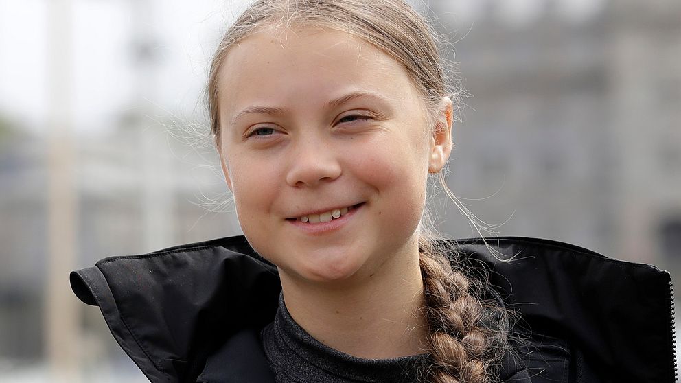 Greta Thunberg i svart jacka och fläta, ler mot kameran.