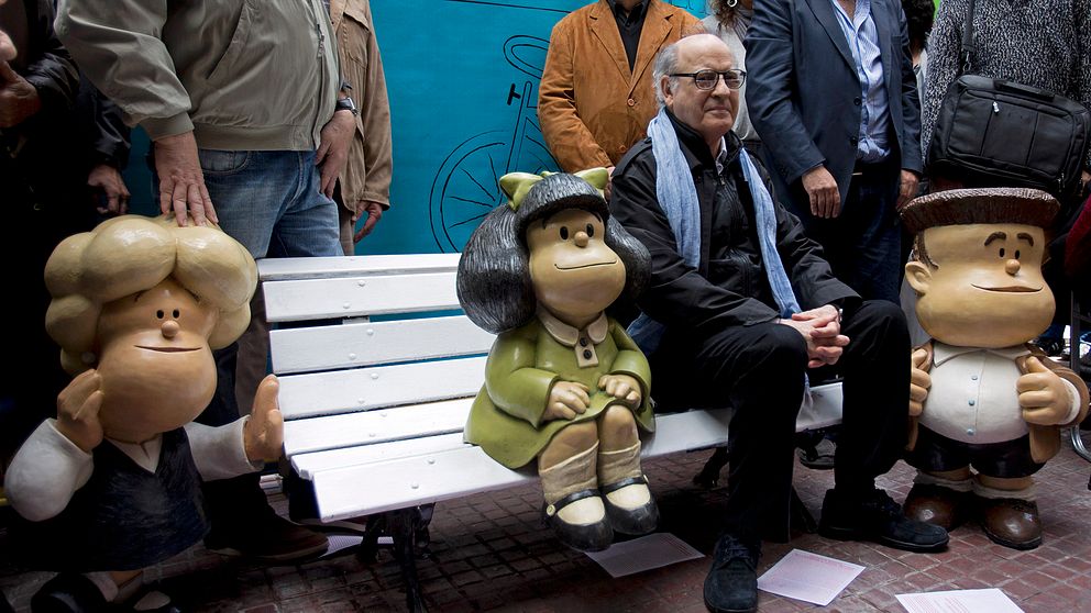 Quino sitter bredvid sin figur Mafalda på en bänk.