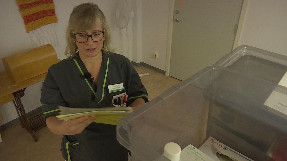 Sjuksköterskan Malin håller i en mapp med instruktioner och står intill en låda med skyddsutrustning.