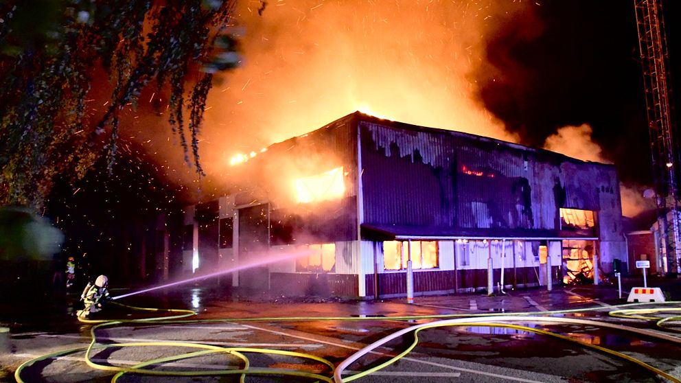 Ett fyrkantigt hus som brinner och en person står på knä med en brandslang som sprutar vatten.