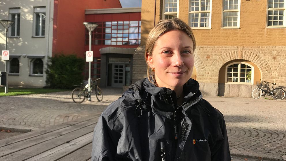 Mikaela Martinsson sitter vid ett bord utanför Strängnäs fritidsgårds entré. Solen skiner och hon har en svart kommunjacka på sig. Hon tittar in i kameran.