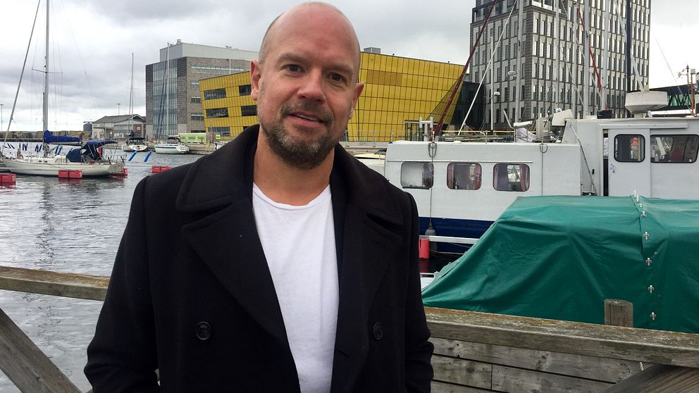 Porträttbild på Pär Israelsson, evenemangschef på Destination Kalmar. Han har på sig en svart rock och vit t-shirt. I bakgrunden syns hav, några båtar samt Linnéuniversitetet.
