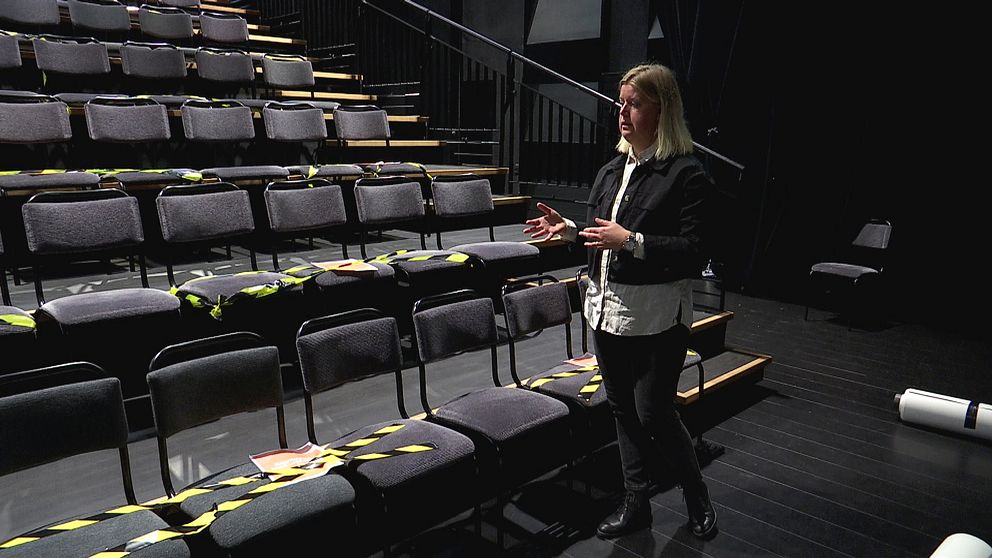 Linda Stenberg, teaterchef på Byteatern i Kalmar står med händerna framför sig och visar hur Byteatern har coronasäkrat sina sittplatser. Linda Stenberg står framför gradängen med svarta stolsrader. En del av stolarna har gul och svart avspärringstejp på sig.