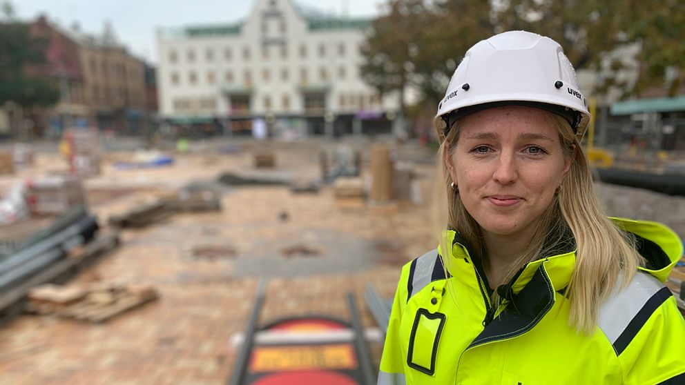 Kvinna i bygghjälm och gul jacka står framför ett torg under uppbyggnad.