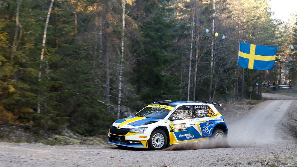 En rallybil på en grusväg. En svensk flagga hänger i bakgrunden.