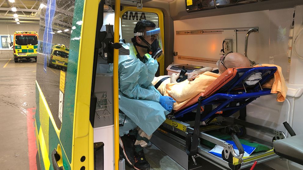 En vårdanställd i skyddsutrustning sitter bredvid en patient på brits i en ambulans.
