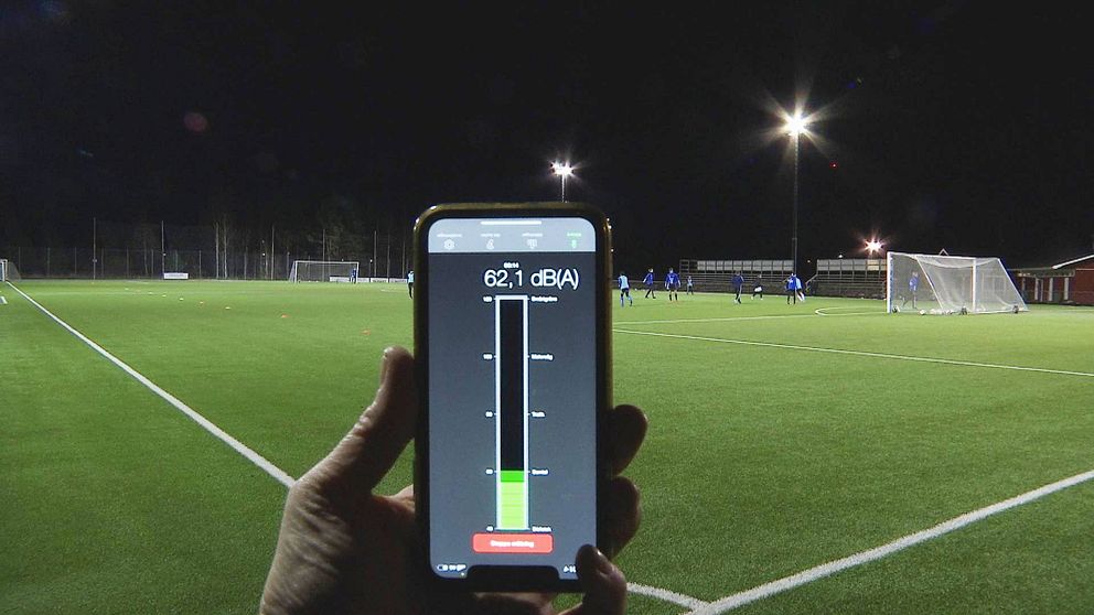 En mobiltelefon där man ser ett mått på ljudnivån hålls upp vid en fotbollsplan där träning pågår.