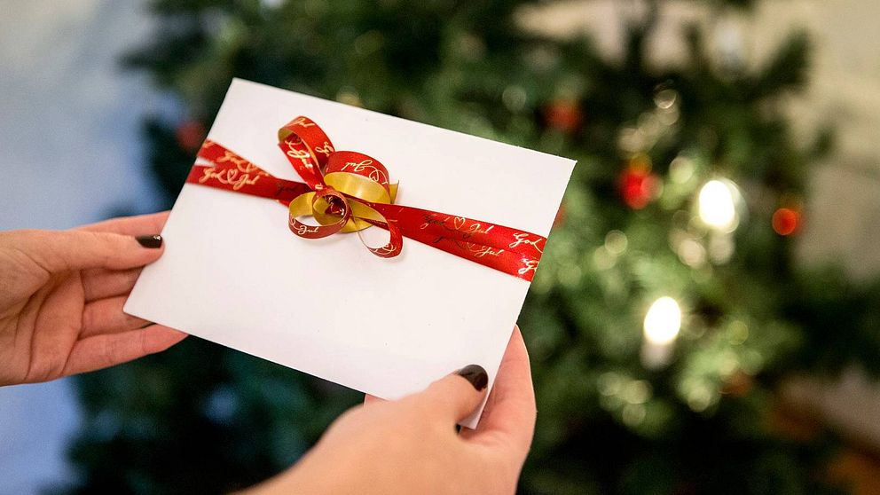 Bild på två händer som håller i ett vitt kuvert med ett rött presentsnöre på. I bakgrunden en suddig julgran.