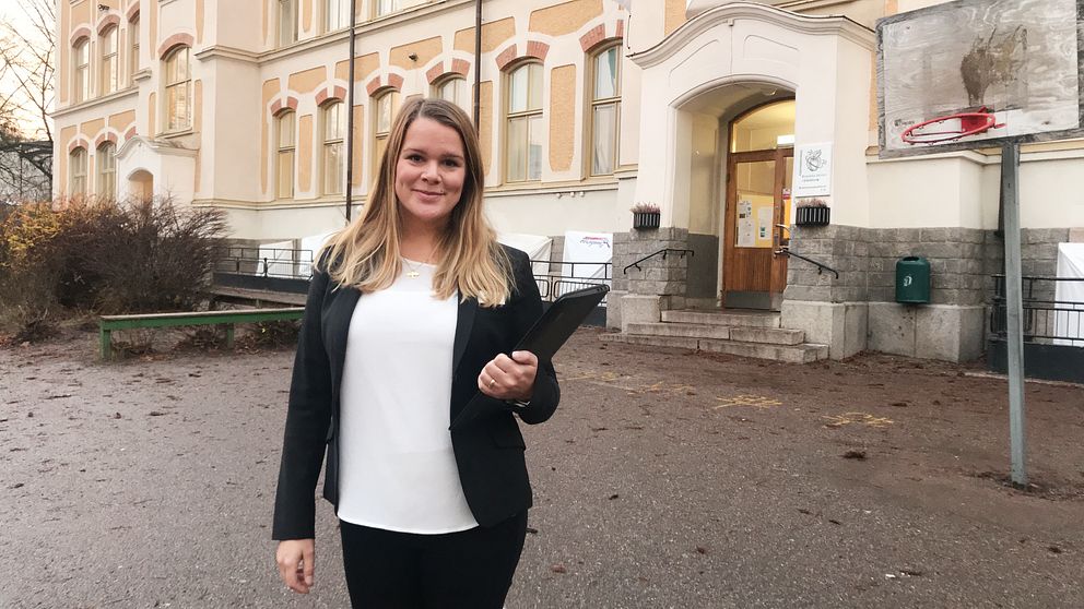 Josefin Korsbäck i svart kavaj står framför Engelska skolan i Nyfors. Hon håller en pärm i ena handen och i bakgrunden syns en basketkorg.