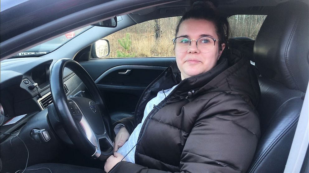 Julia sitter i sin bil, iklädd svart vinterjacka och glasögon.