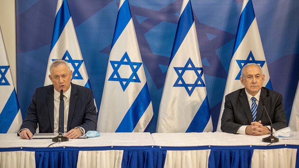 Benny Gantz och Benjamin Netanyahu sitter med två meters avstånd till varandra under en presskonferens med fem israeliska flaggor bakom sig.