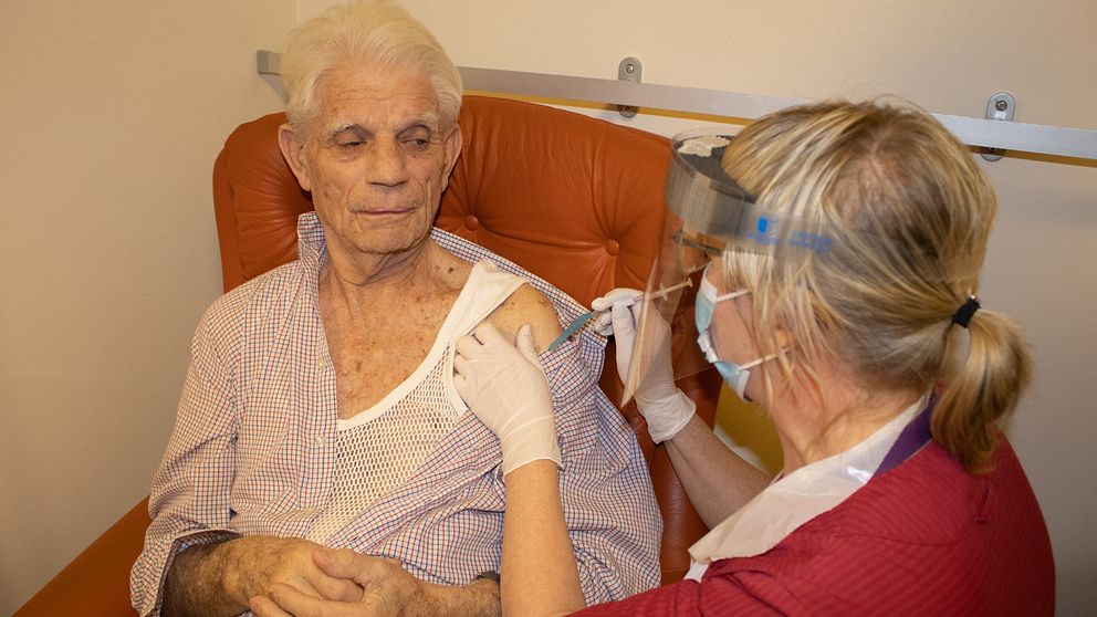 Bilden föreställer 88-årige Rune Rosander. Han sitter i en brun skinnfåtölj med händerna knäppta. Han tittar på sjuksköterskan Eva Yngvesson som injicerar covidvaccin i hans arm.