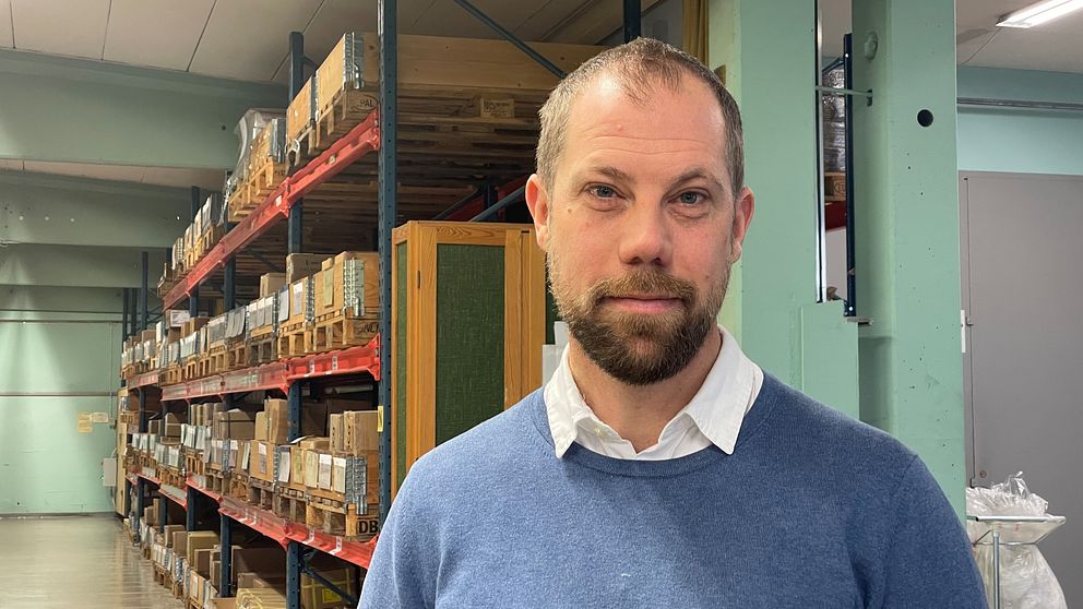 MEDELÅLDERSMAN TITTAR IN I KAMERAN. LAGERGÅNG MED PALLAR I BAKGRUNDEN.