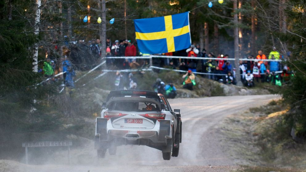 En rallybil i skogen. I bakgrunden publik samt en stor svensk flagga.