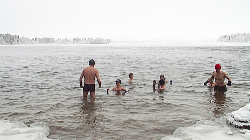 Flera personer som badar ifrån istäckt strand, snöig skog bortanför vattnet