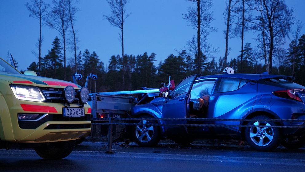 Bilden föreställer en krock utanför Växjö. I förgrunden på vänster sida skymtar ett gult räddningstjänstfordon. Den står bredvid ett vajerräcke. På andra sidan står en blå bil som kolliderat med ett bilsläp. Huven är krossade och airbagen har lösts ut.