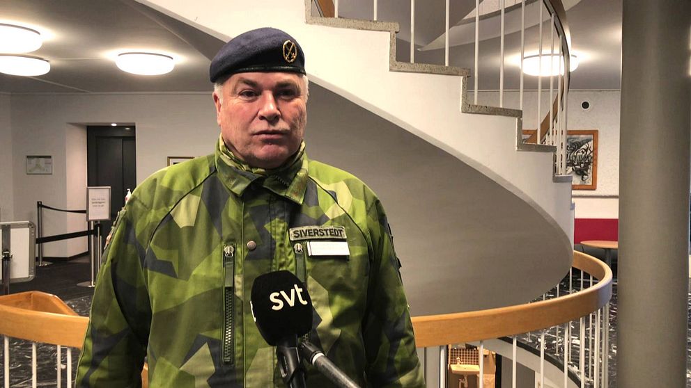 en medelålders man i militärens kamouflagekläder och basker intervjuas framför en trappa
