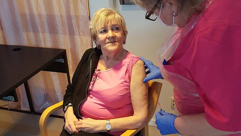 Blond kvinna klädd i rosa tröja och svarta byxor sitter i en stol En annan kvinna står bredvid och ska ge en vaccinationsspruta i vänsterarmen på hon som sitter i stolen.