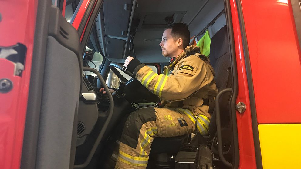 Bilden föreställer en brandman som sitter i en brandbil. Han syns från sidan, från smalbenen och uppåt. Han tittar framåt ut genom rutan och hans ansikte syns i profil.