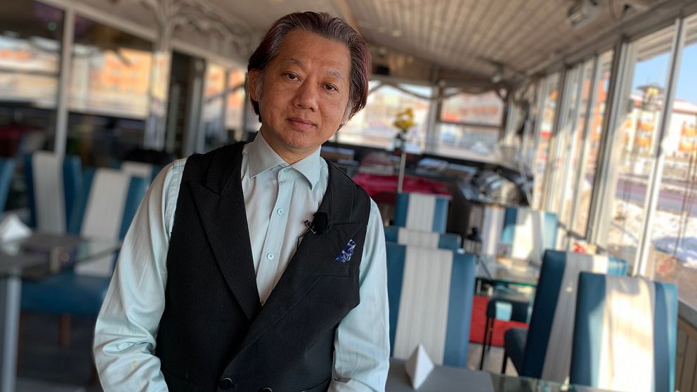 En vietnamesisk medelåldersman står i en tom restaurang. Han syns från midjan och uppåt. Han har iklädd vitskjorta och svart väst. Bakom honom syns blåvita stolar och glasbord med vita servetter. Väggarna består av stora glaspartier.