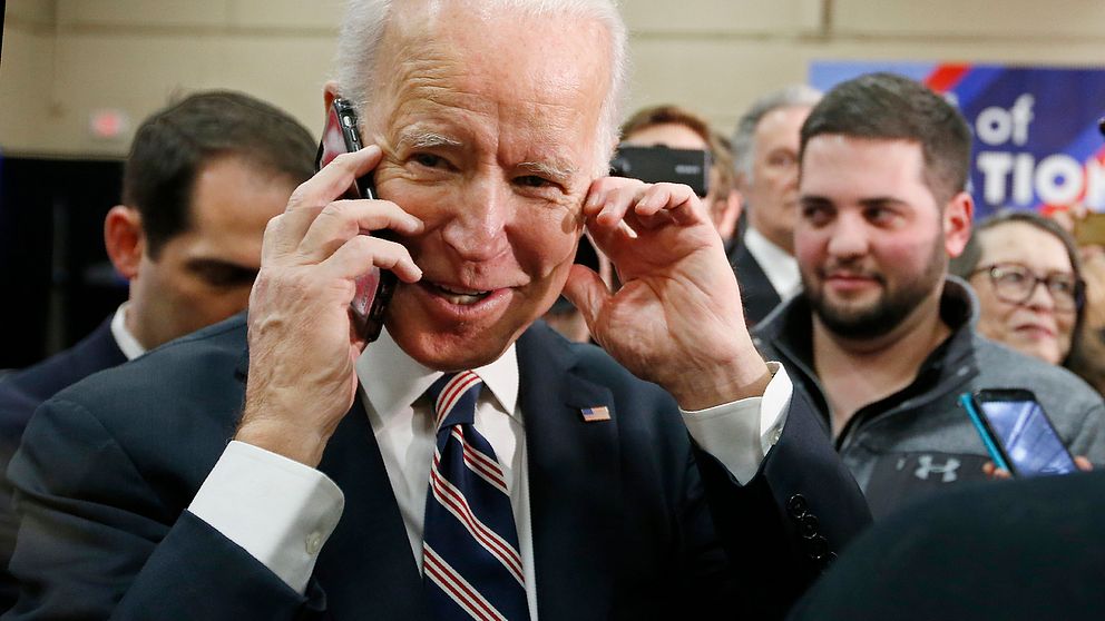 En glad Joe Biden talar i telefon, omgiven av människor.