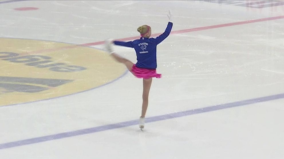 flicka med blont hår blå jacka och rosa kjol gör en piruett på en isrink