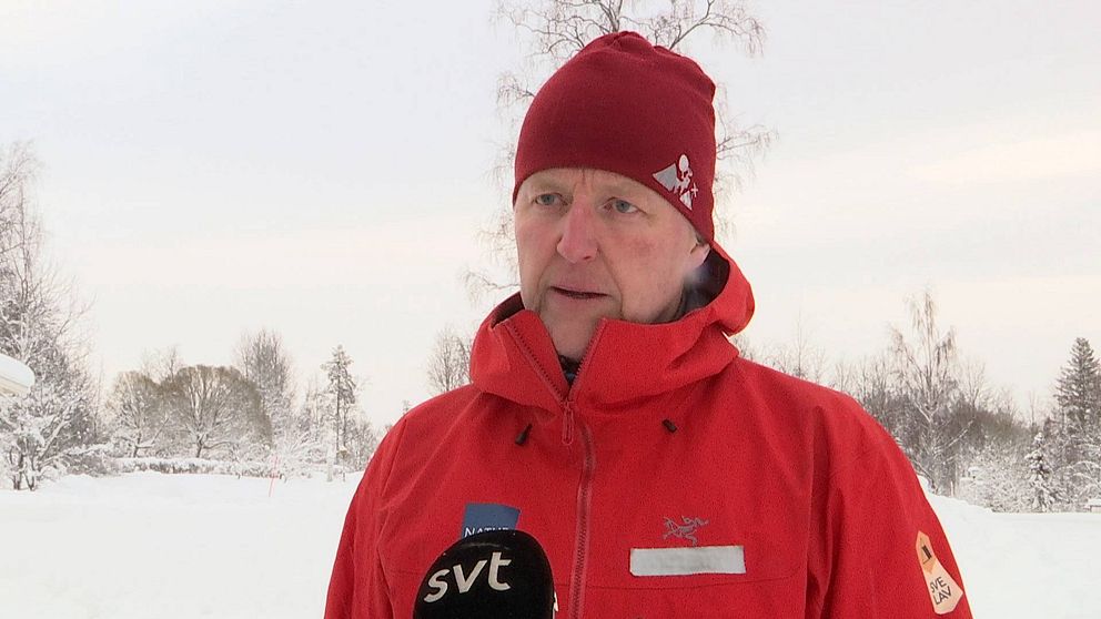Man i röd mössa och röd jacka intervjuas i ett vitt vinterlandskap.