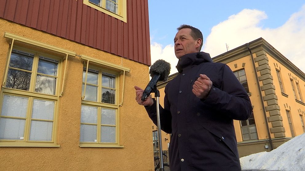 Man i blå jacka står vid ett mikrofonstativ. Tittar åt vänster. Bilden är tagen lite underifrån med två gulröda hus i bakgrunden.