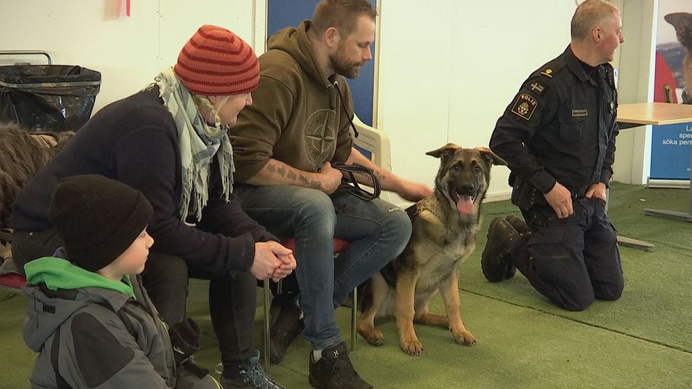 man i polisunuform och ett barn, en kvinna och en man sitter med en ung schäferhund, en man i polisuniform hukar bredvid.