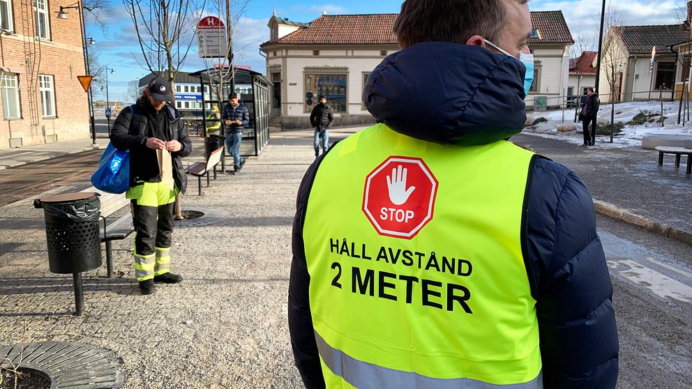 Man i reflexväst på en busshållplats. Han står med ryggen mot kameran. På ryggen står det ”STOP HÅLL AVSTÅND 2 METER”. Han bär munskydd.