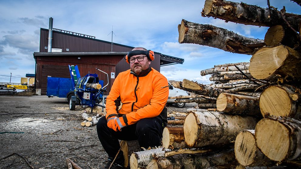 en man i varseljacka och hörselkåpor sitter vid en stapel björkstammar, vedkap och förrådslada i bakgrunden