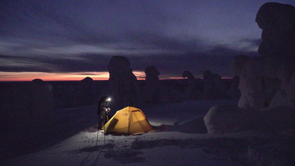 person med pannlampa står vid tält nattetid, träd inbakade i snö skymtar i mörkret