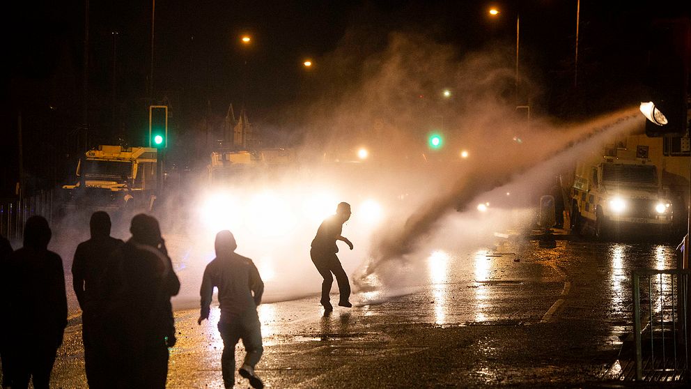 En vattenkanon skjuter en sotr stråle vatten mot svartklädda demonstranter i motljus. Våldsamheter bröt ut under torsdagen i västra Belfast när nationalistiska ungdomar attackerade polisen, som skingrade massorna med vattenkanoner.