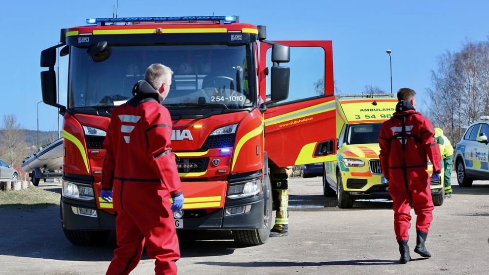 Två personer i dykarkläder sedda bakifrån framför en stor brandbil, en ambulans och en polisbil.