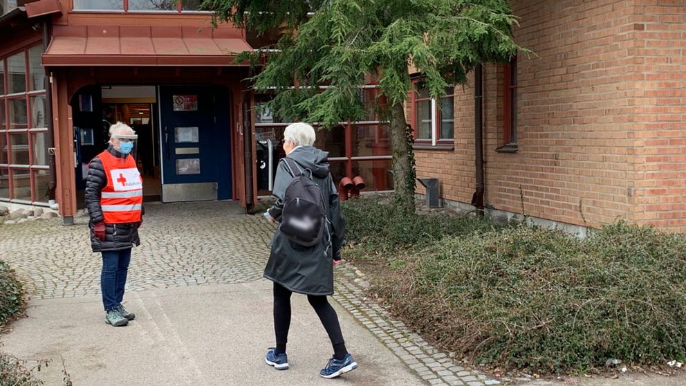 En äldre kvinna är på väg in i Kalmars vaccinationscentral. Utanför möts hon av en annan kvinna som arbetar som funktionär. Hon har skyddsutrustning och reflexväst.