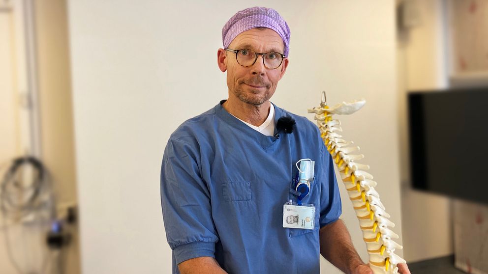 Harald Zetterquist i blå sjukhusklädsel står bredvid en modell av en ryggrad.