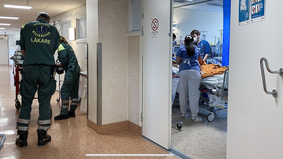 personal från ambulanshelikopter rullar in en brits i korridoren medan intensivvårdspersonal ses vårda en coronapatient i ett rum