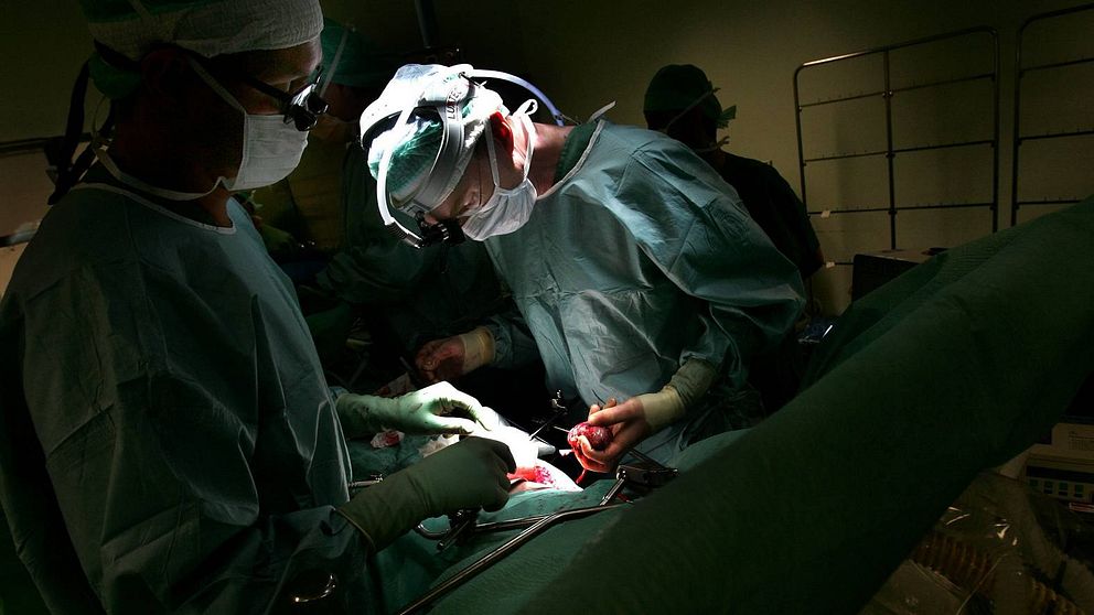 Grönklädda personer med munskydd står i ett mörkt rum och utför en operation.