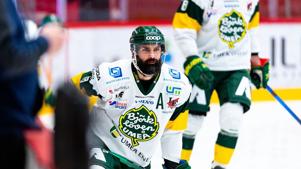 En man i grön ishockeyhjälm och tätt skägg står på knä på en isrink med fokuserad blick.