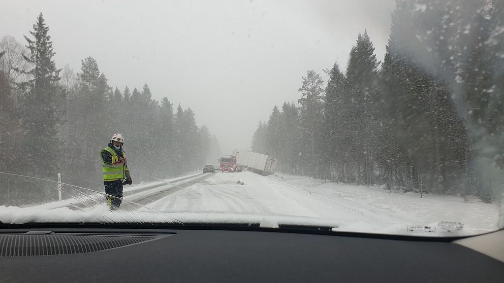 En snöig väg med en räddningstjänstpersonal i förgrunden och en lastbil med ett släp i diket på bakgrunden.