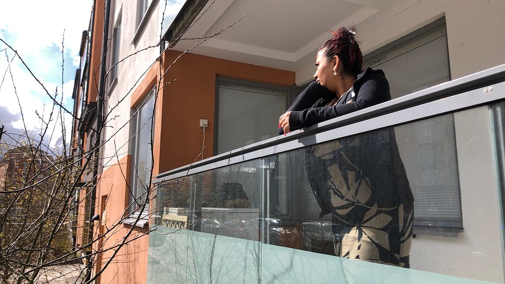 Elsa Olah står på sin balkong med ena armen på balkongräcket och tittar till vänster i bild mot platsen där explosionen inträffade. Till vänster i bild ser man några trädgrenar och husfasaden som är ljus och röd.