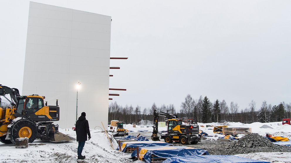 Två traktorer som jobbar med att bygga upp en ny fabrik. En stor vit vägg och en människa framför.