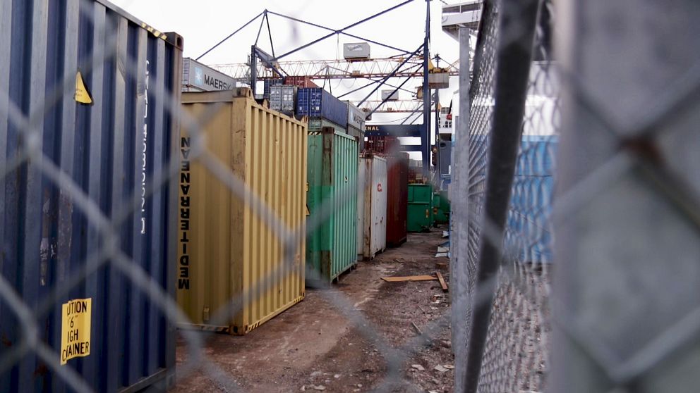 containrar på rad ses genom ett staket i hamnområdet, kranar i bakgrunden