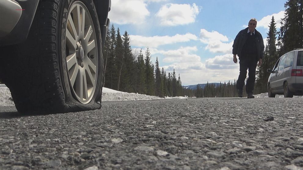 närbild på punkterat däck på en bil längs vägen, en person går i bakgrunden från en annan bil