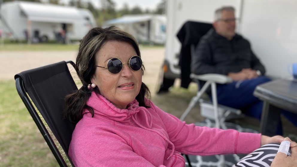 en medlelålders kvinna i solglasögon och rosa luvtröja sitter i en campingstol utanför husvagn