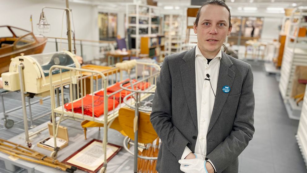 Jean-Pierre Salkvist står intill några sjukhusföremål från 1900-talet, bland annat en säng, ett par kryckor och en docka.