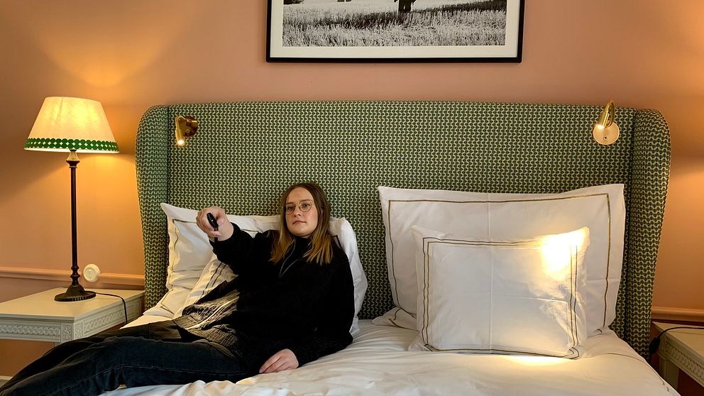 Reporter lutar sig tillbaka i hotellsängen och håller en fjärrkontroll i handen. Till vänster i bild syns en lampa, väggarna är i persikofärg och över sängen hänger en tavla.