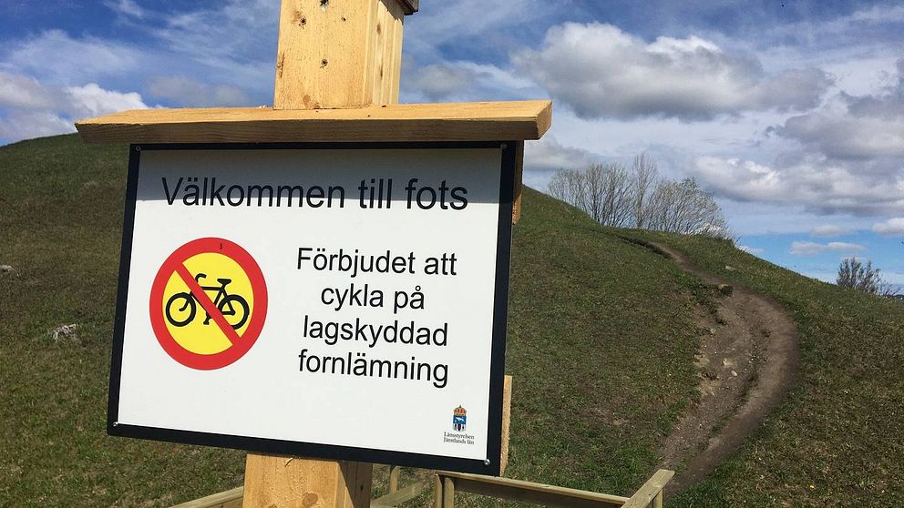 Skylt med texten ”Välkommen till fots. Förbjudet att cykla på lagskyddad fornlämning”. I bakgrunden en rejäl stig uppför en kulle.