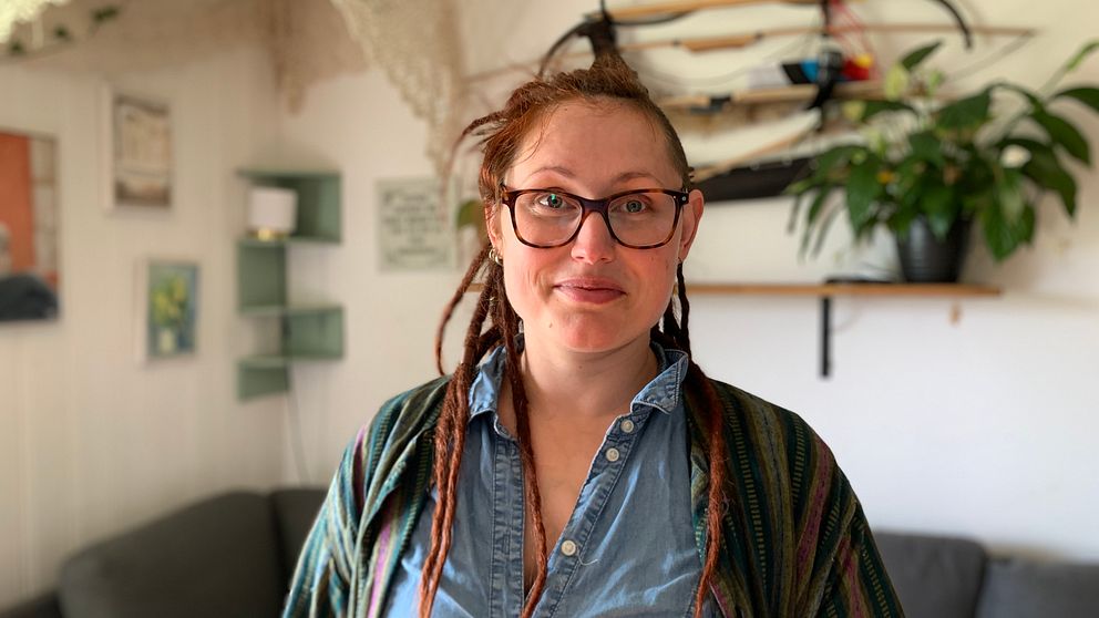 Tanja Östberg har röda dreadlocks och glasögon. Tanja har en jeanblus och en cardigan på sig. Hon står i sitt vardagsrum framför soffan och poserar. I bakgrunden hänger några pilbågar på väggen.