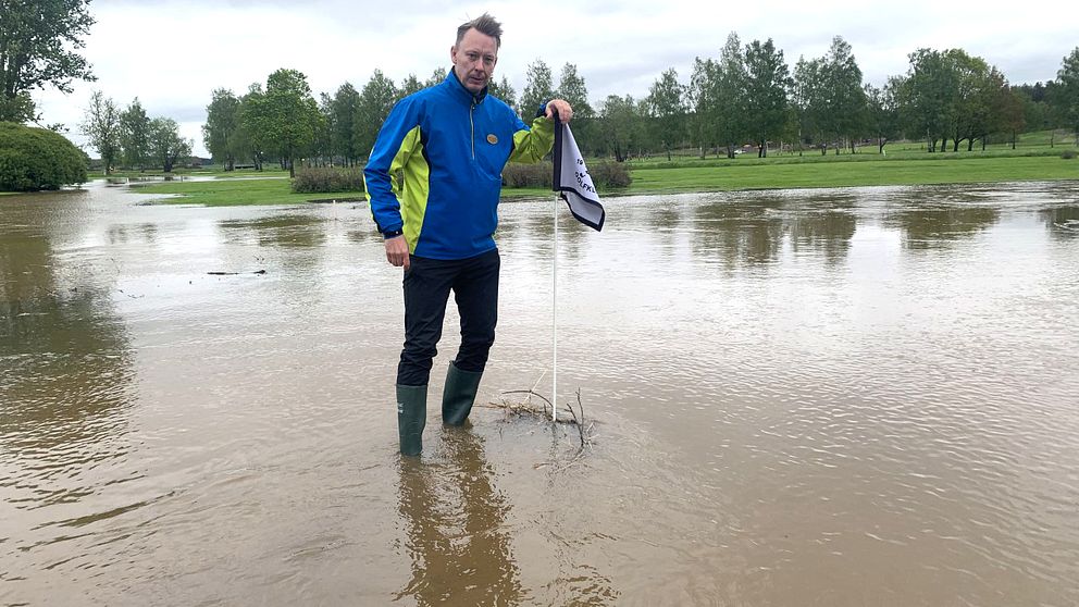 Den stövelbeklädde klubbchefen Richard Svalling står på den översvämmade golfbanan med vatten över anklarna.